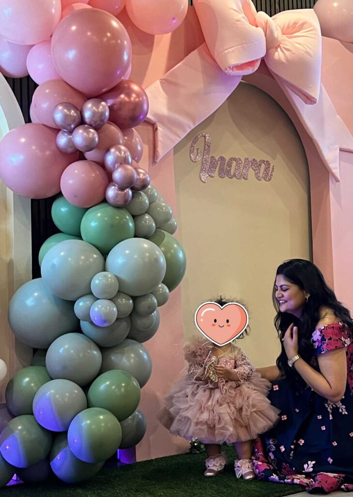 Inara's 1st birthday: pastel balloon arch, girl in pink dress.